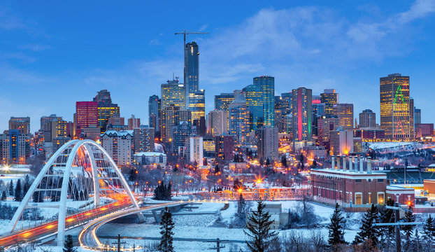 Snowy city skyline at dusk with illuminated skyscrapers and a curved arch bridge in the foreground.