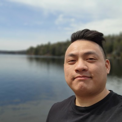 Selfie of a man with black hair wearing a black shirt, beside a calm lake with trees in the background.