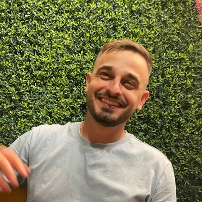 Smiling man with short light brown hair and a beard, wearing a light gray T-shirt, standing in front of a dense green hedge.