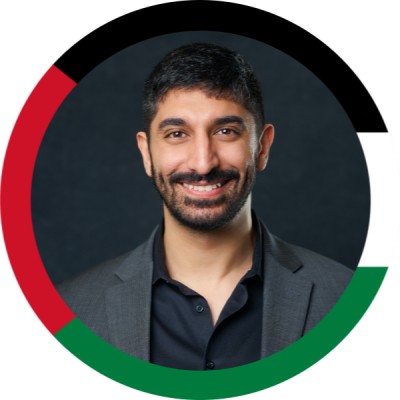 Smiling man in a dark suit and open-cneck shirt, head-and-shoulders portrait, encircled by a multicolor ring in red, black, and green.