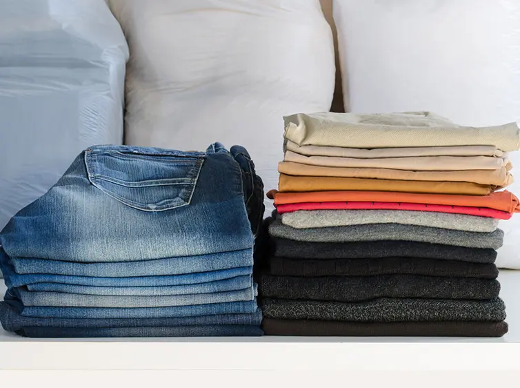 Folded blue jeans on the left and a stack of folded clothes in beige, orange, gray, and black on the right on a shelf.