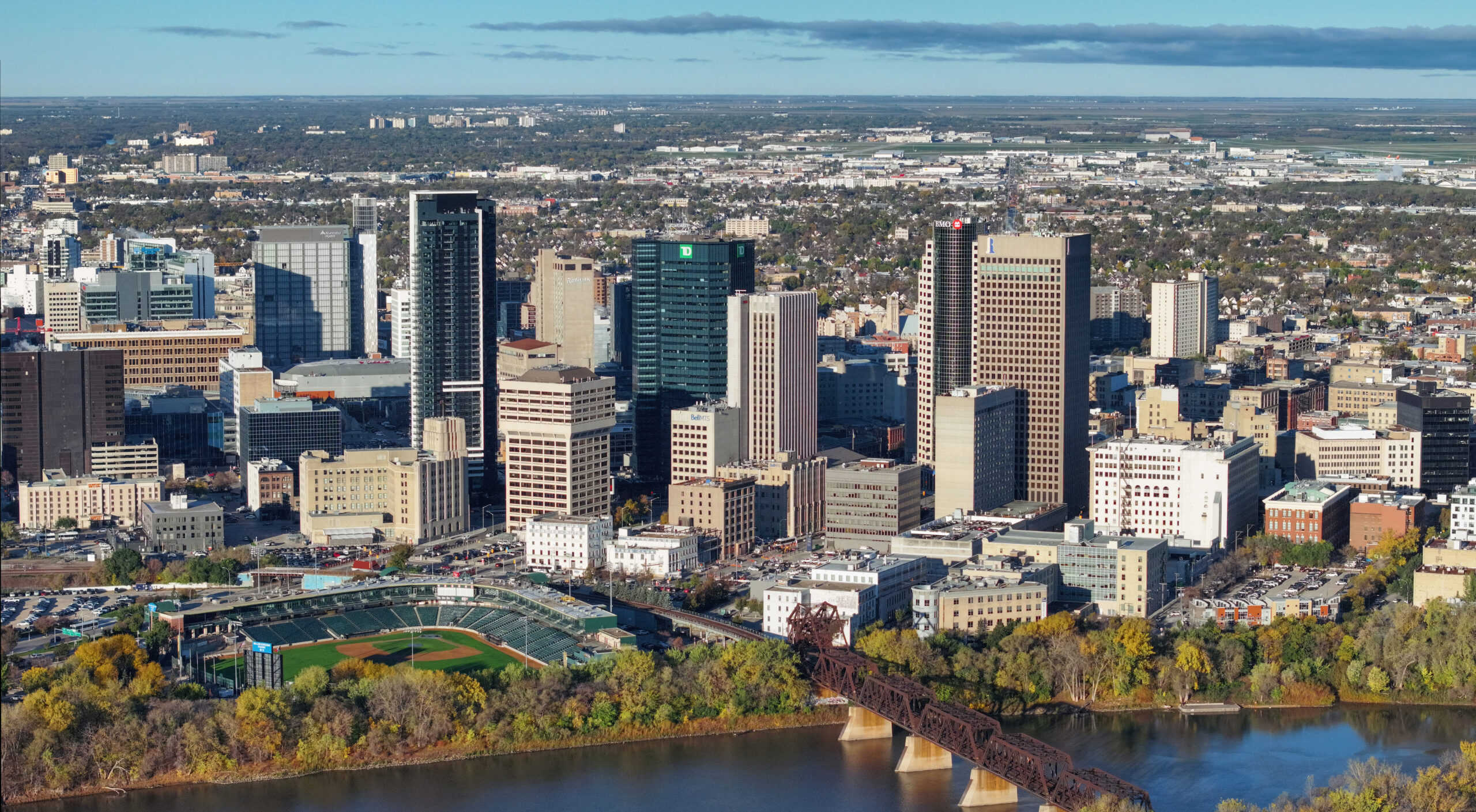 Aerial view of a downtown skyline along a river, with tall office buildings, a bridge crossing the water, and a baseball stadium at the bottom left.